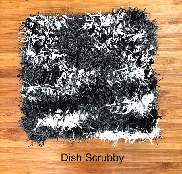 Top-down detailed view of a square handmade crocheted dish scrubby made with fuzzy, textured eyelash yarn in black, gray, and white stripes, resting flat on a wooden surface.