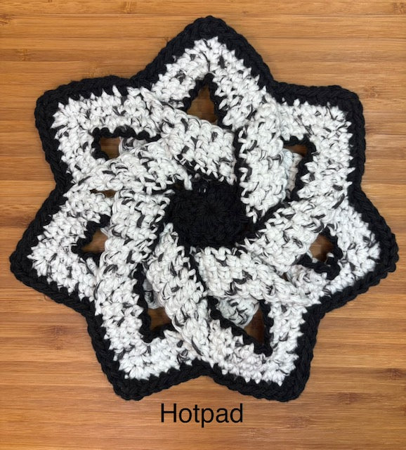 Top-down view of a handmade crocheted star-shaped hot pad trivet with an intricate interlocking design in black and speckled white yarn, sitting flat on a smooth wooden cutting board.