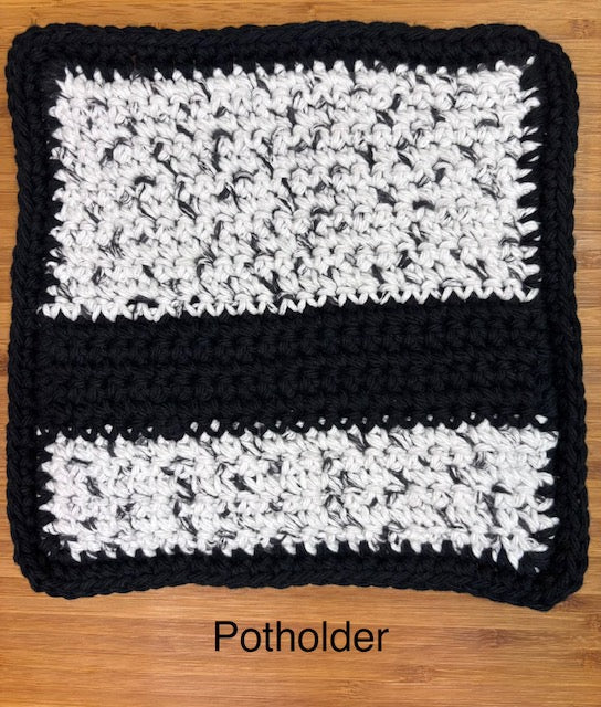 Close-up of a handmade crocheted square potholder featuring thick black borders, solid black stripe, and speckled black and white yarn panels, displayed flat on a wooden surface.