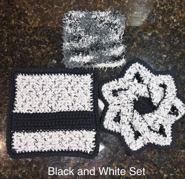 Three-piece handmade crocheted kitchen set in black, white, and gray, featuring a swirling star-shaped hot pad, a thick striped square potholder, and a textured fuzzy square dish scrubby.
