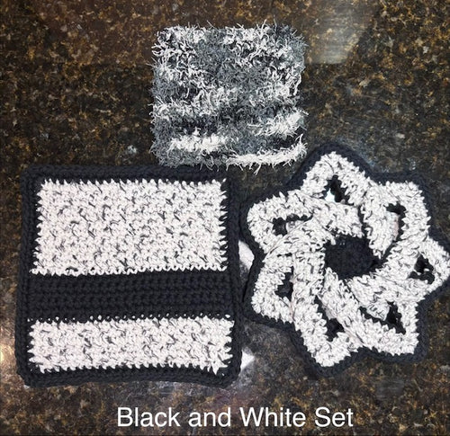 Three-piece handmade crocheted kitchen set in black, white, and gray, featuring a swirling star-shaped hot pad, a thick striped square potholder, and a textured fuzzy square dish scrubby.