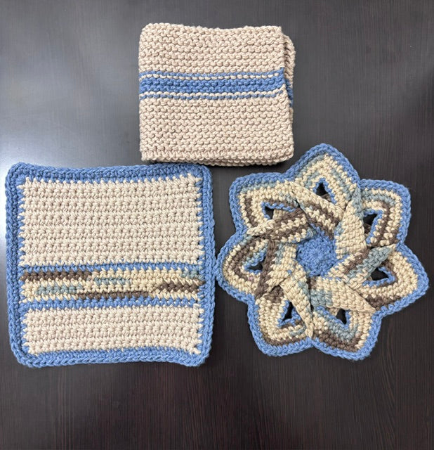 Three-piece handmade crocheted kitchen set in soft denim blue, beige, and brown earth tones, featuring a folded striped dishcloth, a square potholder, and a matching star-shaped hot pad trivet.