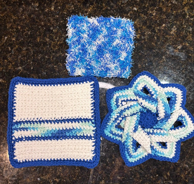Three-piece handmade crocheted kitchen set in vibrant royal blue and aqua tones, featuring a swirling star-shaped hot pad trivet, a square striped potholder, and a matching fuzzy square dish scrubby.