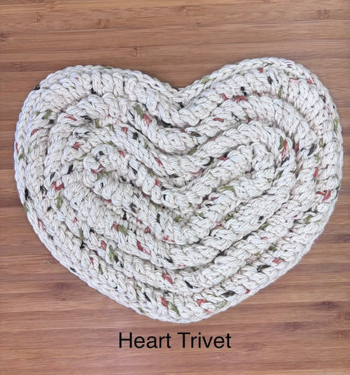Cream-colored handmade crocheted heart trivet featuring a braided spiral pattern with subtle red, green, and gold holiday flecks, displayed flat on a wooden cutting board.