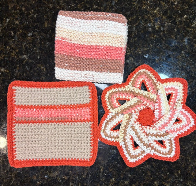 Three-piece handmade crocheted kitchen set in warm terracotta tones, including a square dishcloth, a striped square potholder, and a swirling star-shaped hot pad trivet.