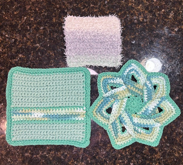 Three-piece handmade crocheted kitchen set in sage green tones, featuring a swirling star-shaped hot pad, a square potholder with blue and green stripes, and a textured square dish scrubby.